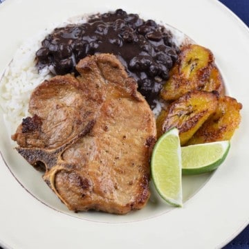 A thin cut, bone in pork chop served with white rice, black beans and fried sweet plantains on a white plate