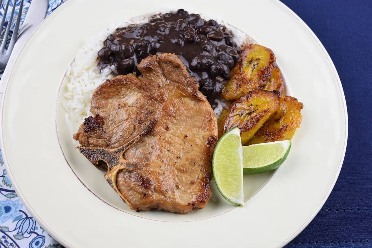 A thin cut, bone in pork chop served with white rice, black beans and fried sweet plantains on a white plate