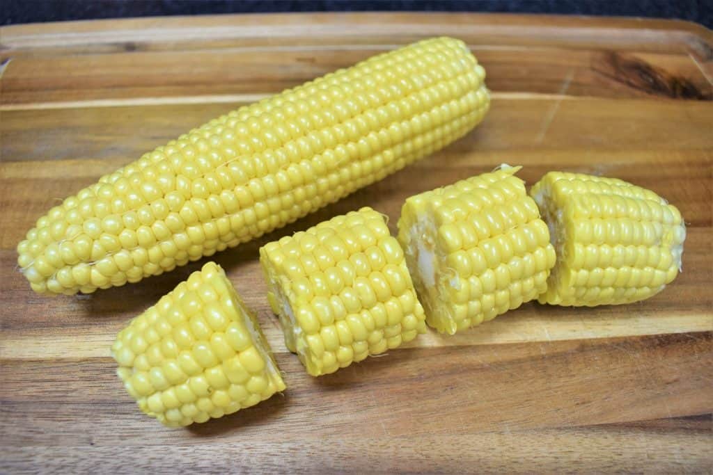 Two corn cobs, one of which as been cut into four pieces, displayed on a wood cutting board.