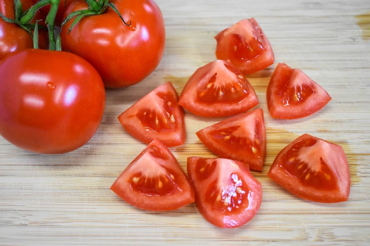 One cut tomatoes on a wood cutting board. With tomatoes whole behind them.