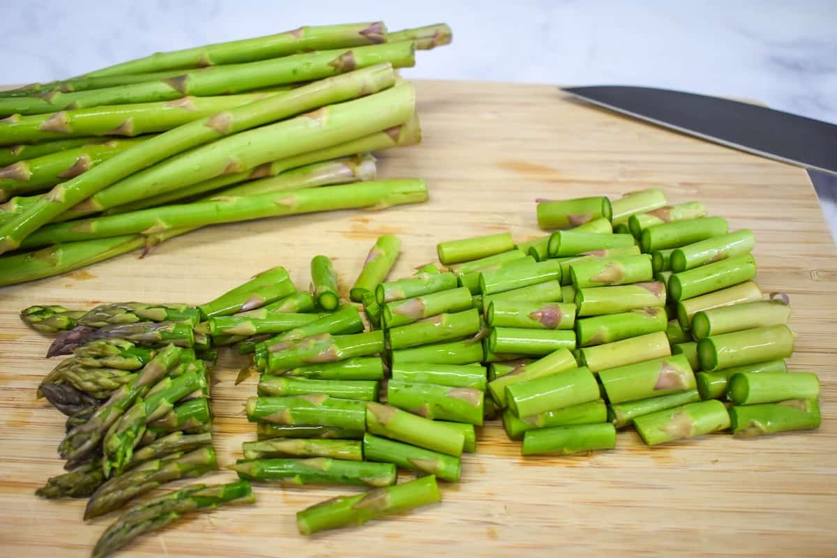 A bunch of asparagus spears cut into one inch pieces on a wood cutting board. The is an uncut bunch in the background.