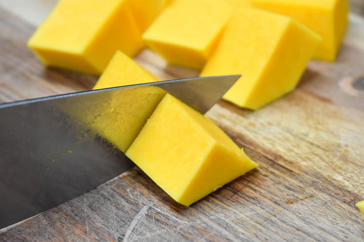 An image of peeled butternut squash being cut into 1 inch pieces on a wood cutting board.