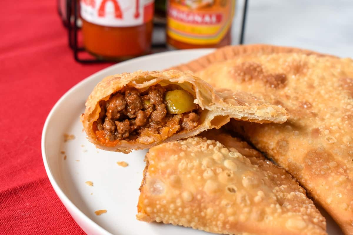 An picture of an empanada cut in half so that the filling is exposed served on a white plate on a red linen with bottles of hot sauce in the background.