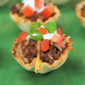 A small fillo dough cup filled with taco meat and topped with cheese, tomatoes, sour cream and tomatoes.