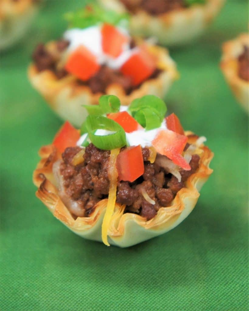 A small fillo dough cup filled with taco meat and topped with cheese, tomatoes, sour cream and tomatoes.