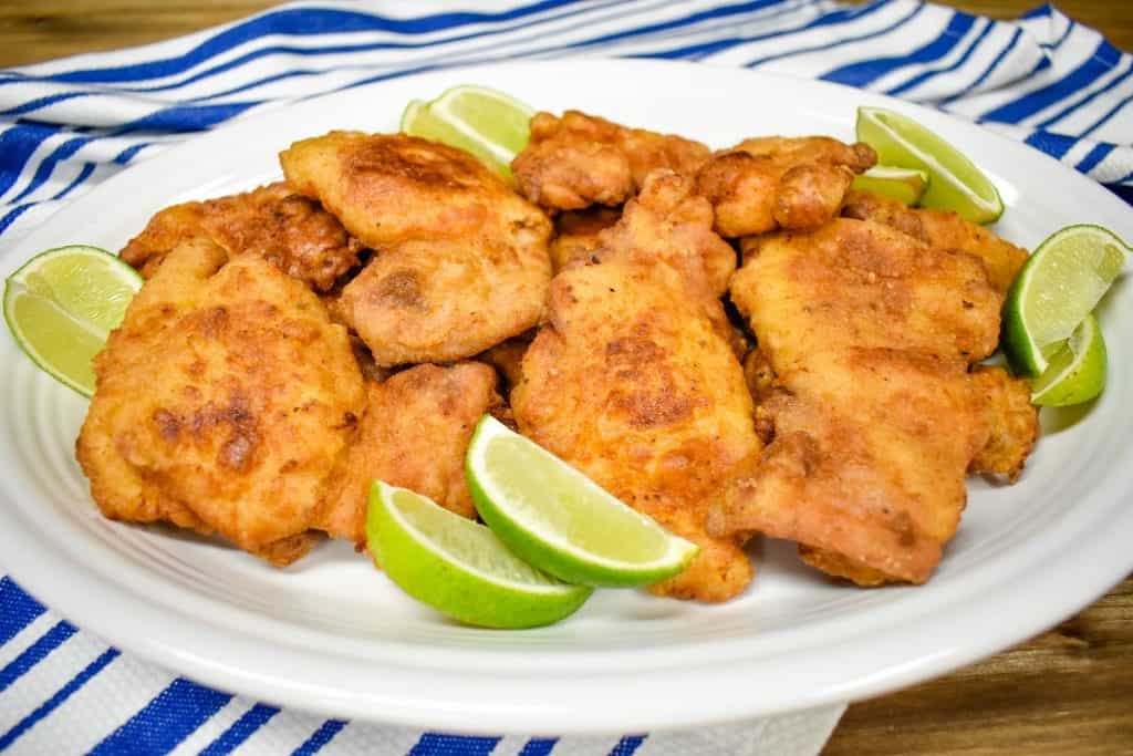 Fried chicken thighs arranged on a large white platter on a blue and white striped cloth and garnished with lime wedges.