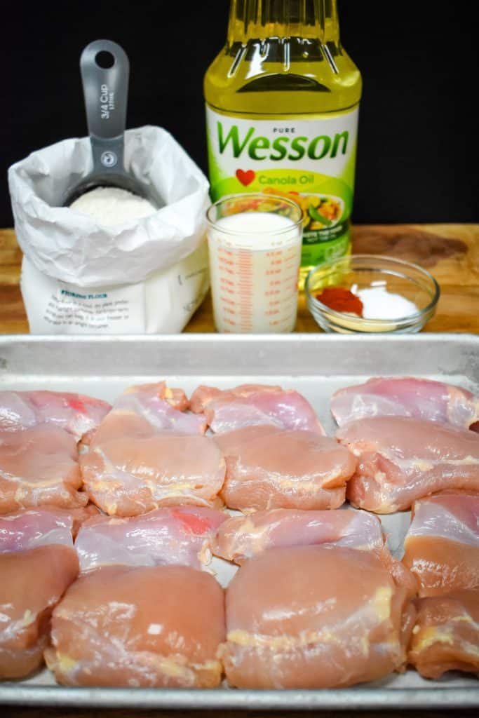 The ingredients for the fried chicken thighs, raw chicken thighs, flour, milk, seasoning and oil displayed on a wood cutting board.