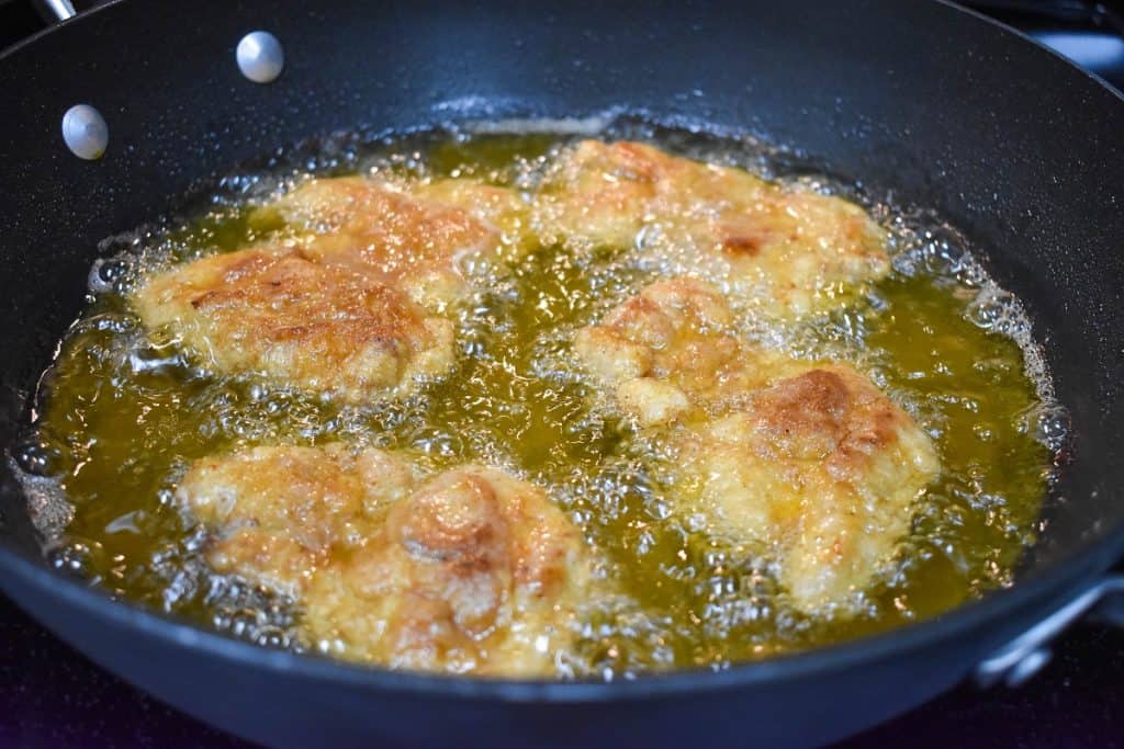 Four pieces of chicken thighs frying in oil in a dark gray skillet.
