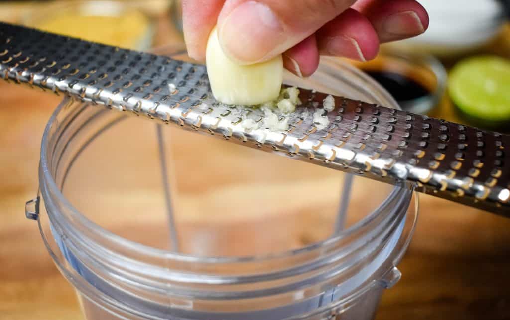 A clove of garlic being grated on a microplane into a jar.