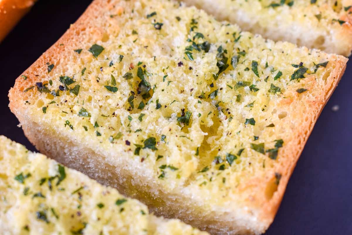 A close up image of a piece of homemade garlic bread displayed on a dark cutting board.