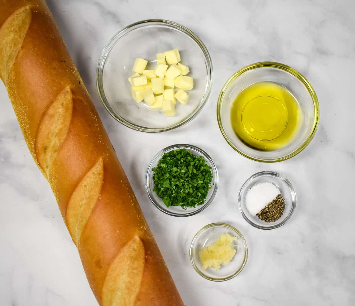 The ingredients for the garlic bread prepped and arranged in glass bowls on a white table with a whole loaf of bread on the left side.