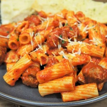 Rigatoni pasta tossed with Italian sausage pasta sauce served on a gray plate with garlic bread in the background.