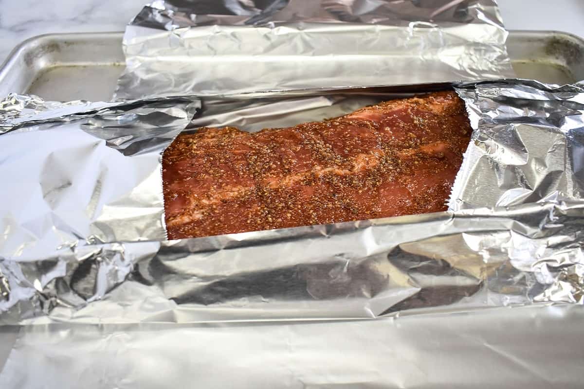 Part of the seasoned rib rack partially wrapped with foil and set on a baking sheet.