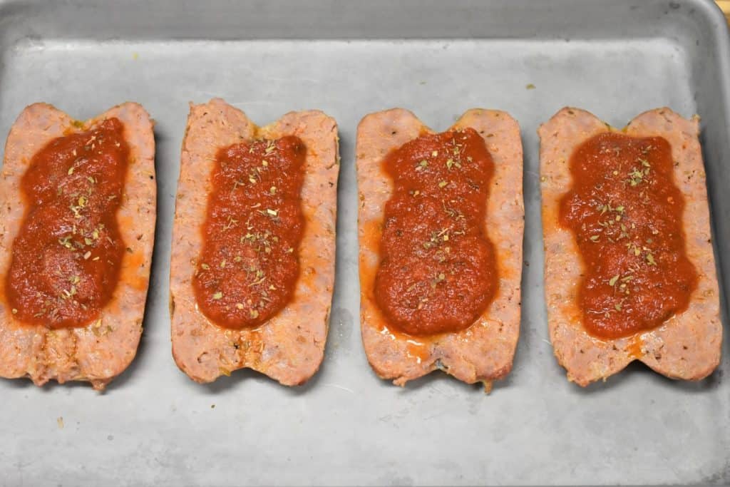 Four butterflied Italian sausages butterflied on a metal baking sheet and topped with tomato sauce and a sprinkle of oregano and garlic powder