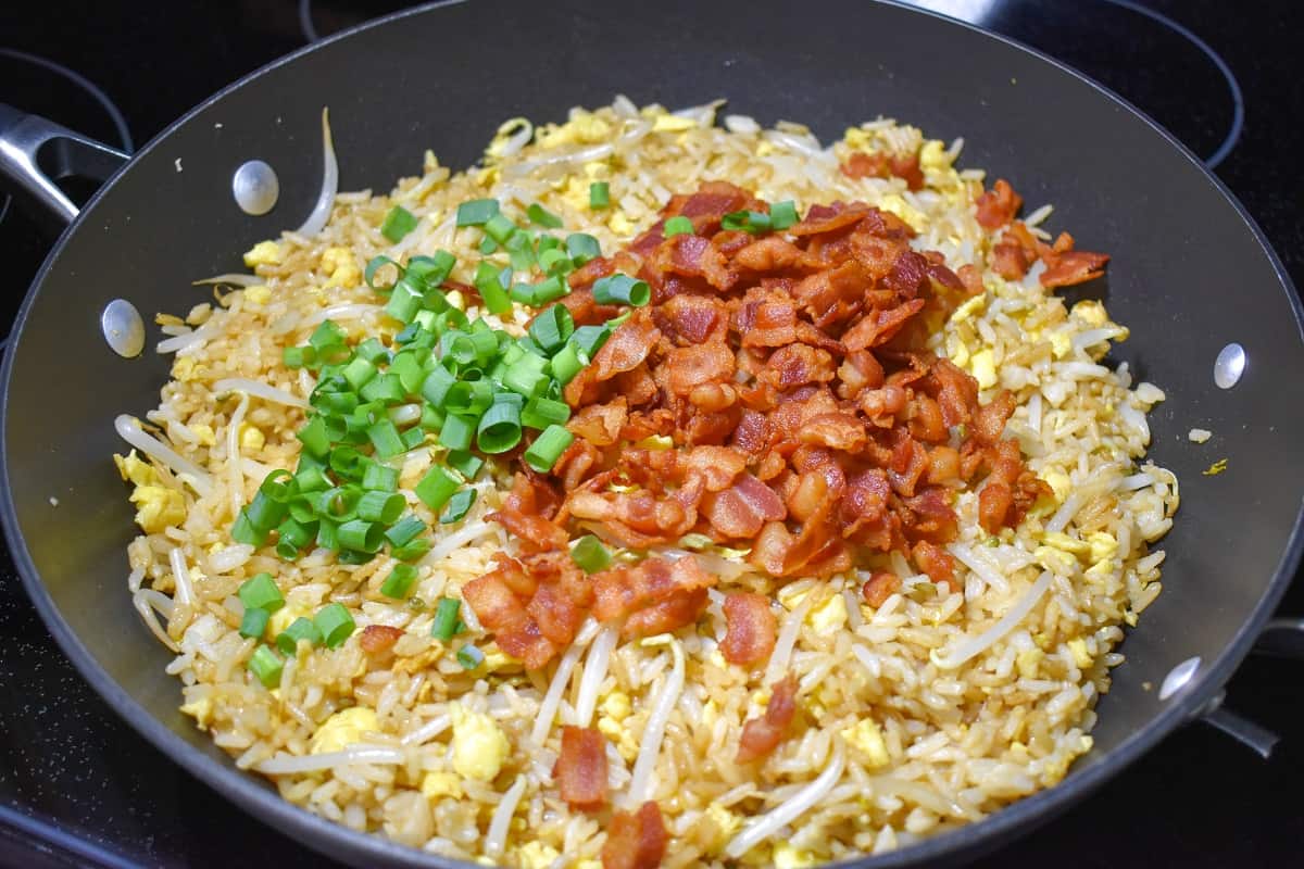 The cooked bacon and green onions on top of the fried rice mixture in a large, black skillet.
