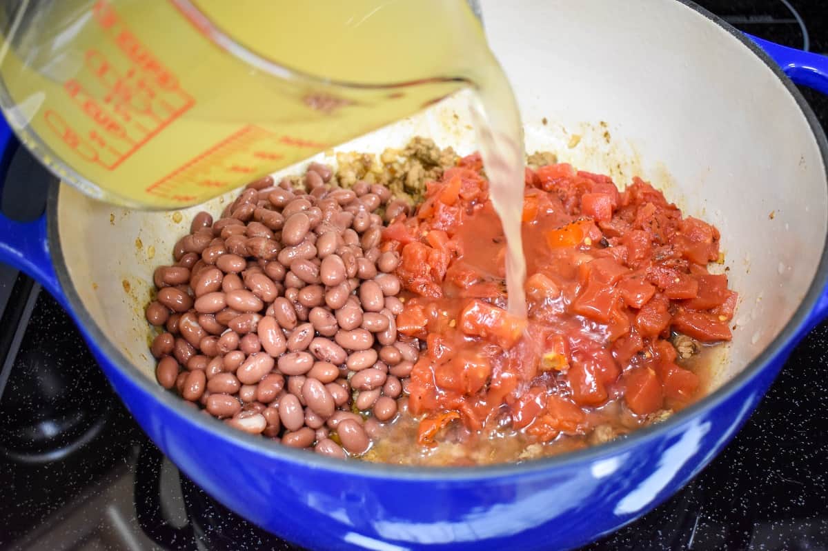 Chicken broth being added to diced tomatoes and red beans in a large blue pot with an off-white interior.