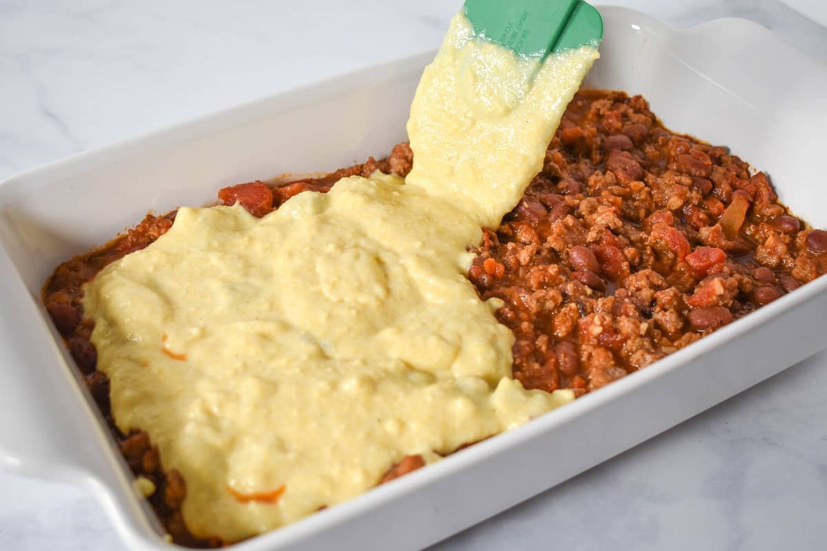 The chili in a white casserole dish with cornbread mix being added to the top.