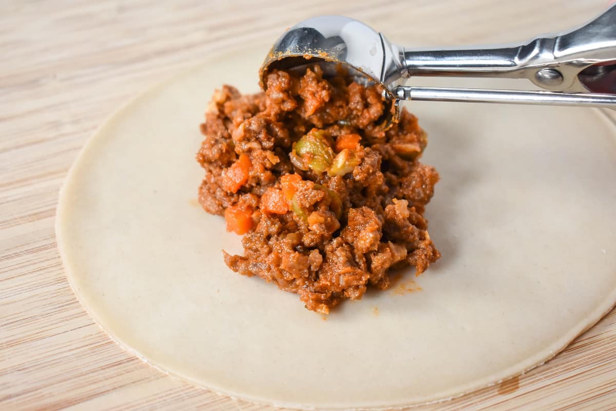 A small scoop adding picadillo filling to the middle of a dough disk.