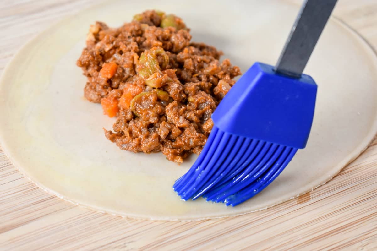 A blue silicone pastry brush applying a thin layer of egg around the empanada dough that has beef filling in the middle.