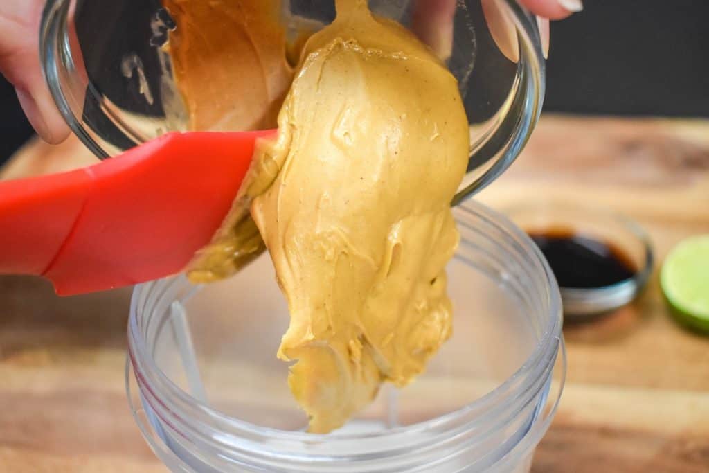 Peanut butter being added to a plastic blender jar using a red, rubber spatula.