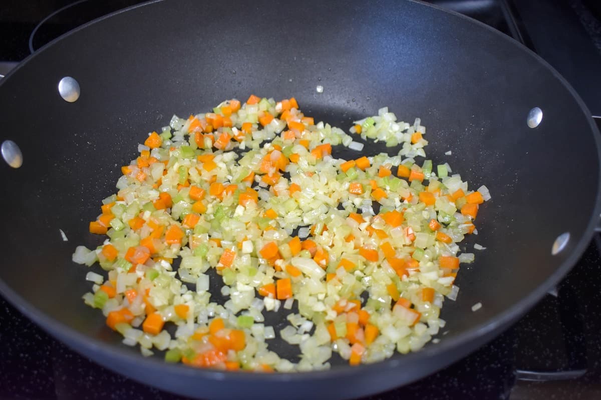 An image of onions, carrots, celery, and garlic cooking in a large, black skillet.