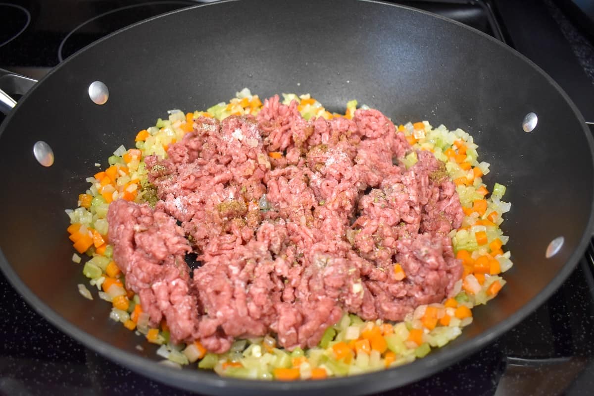 An image of ground beef added to a layer of vegetables in a large, deep skillet.