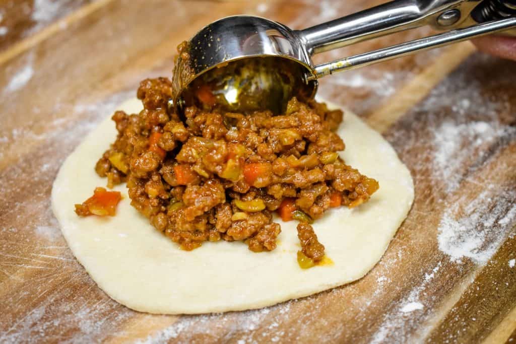 A small metal scoop adding cooked ground beef to a rolled out biscuit round on a floured wood cutting board.