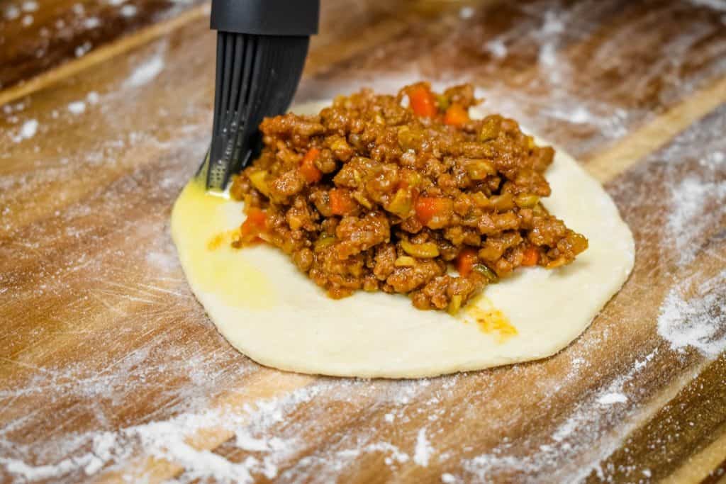 A dark gray silicone brush spreading egg around the biscuit round that has a mound of ground beef filling in the middle. 