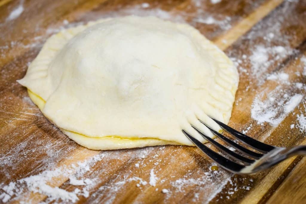 A fork crimping the edge of a ground beef stuffed biscuit, displayed on a floured wood cutting board.