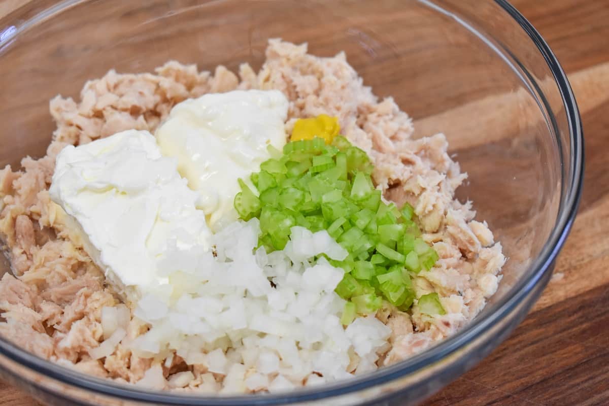 Tuna, mayonnaise, mustard, cream cheese, celery and onions in a large glass bowl before being stirred.