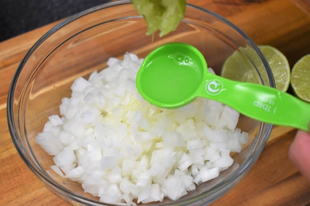 A green tablespoon pouring lime juice on diced white onions in a clear bowl.