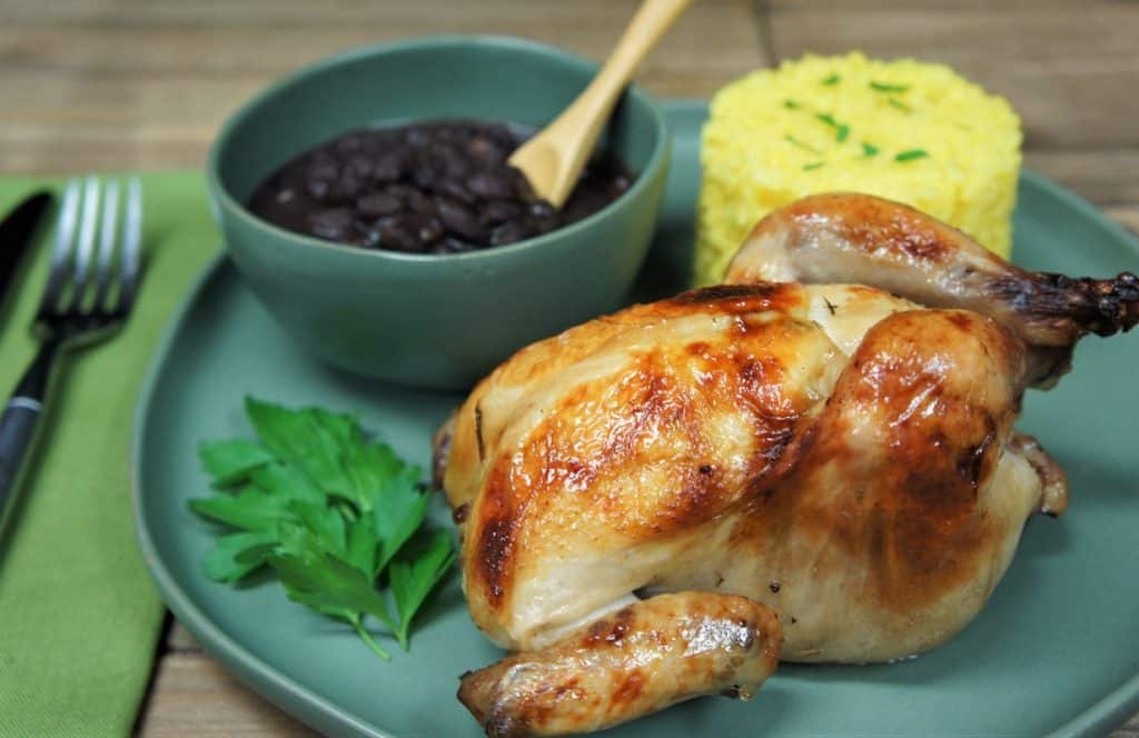 A whole Cornish hen served on a large green plate with yellow rice and a bowl of black beans in the background.
