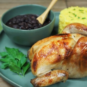 A whole Cornish hen served on a large green plate with yellow rice and a bowl of black beans in the background.