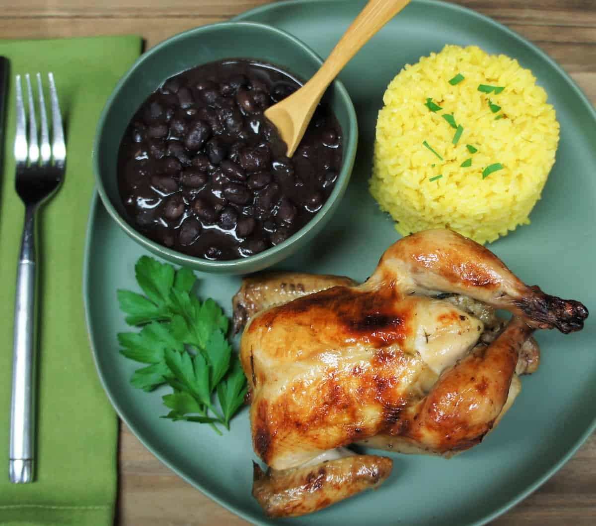 A whole Cornish hen served on a green plate with yellow rice and black beans.