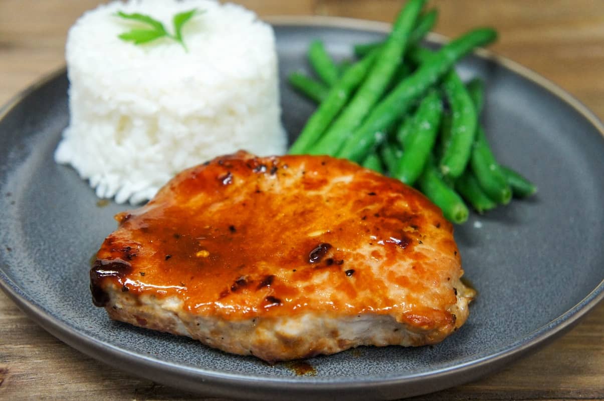 The orange glazed pork chop served on a gray plate with white rice and green beans.