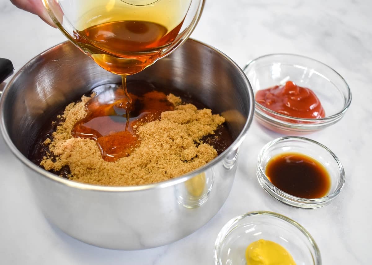 A small sauce pan with the barbecue sauce, brown sugar and the honey being added. The rest of the ingredients for the sauce are next to it in small glass bowls.