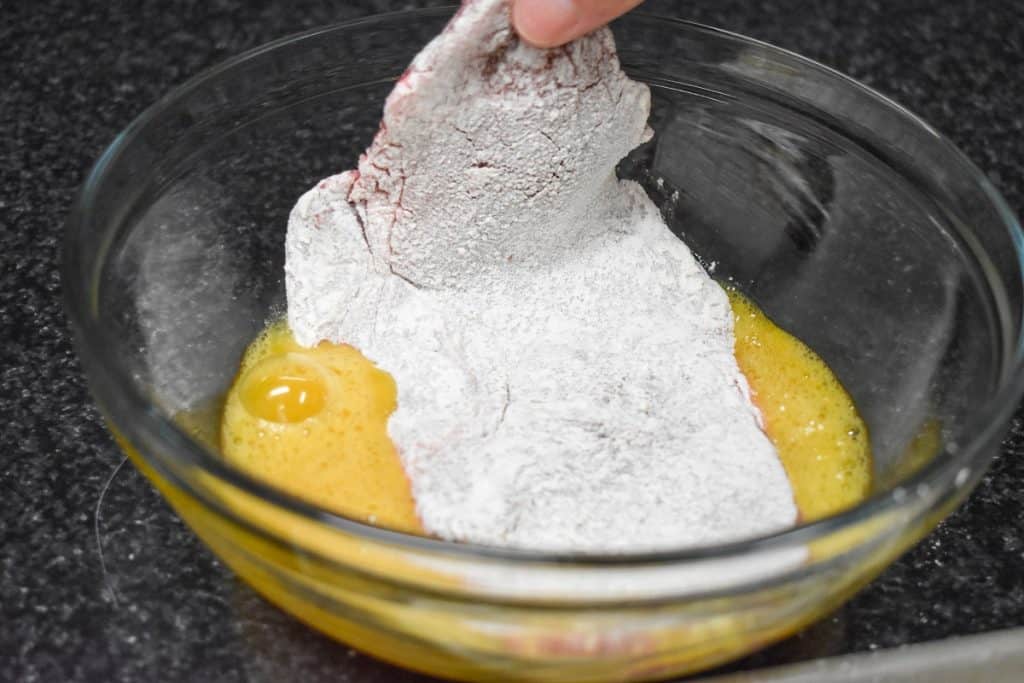 A large, thin steak that's coated in flour and being dipped into a bowl with egg.