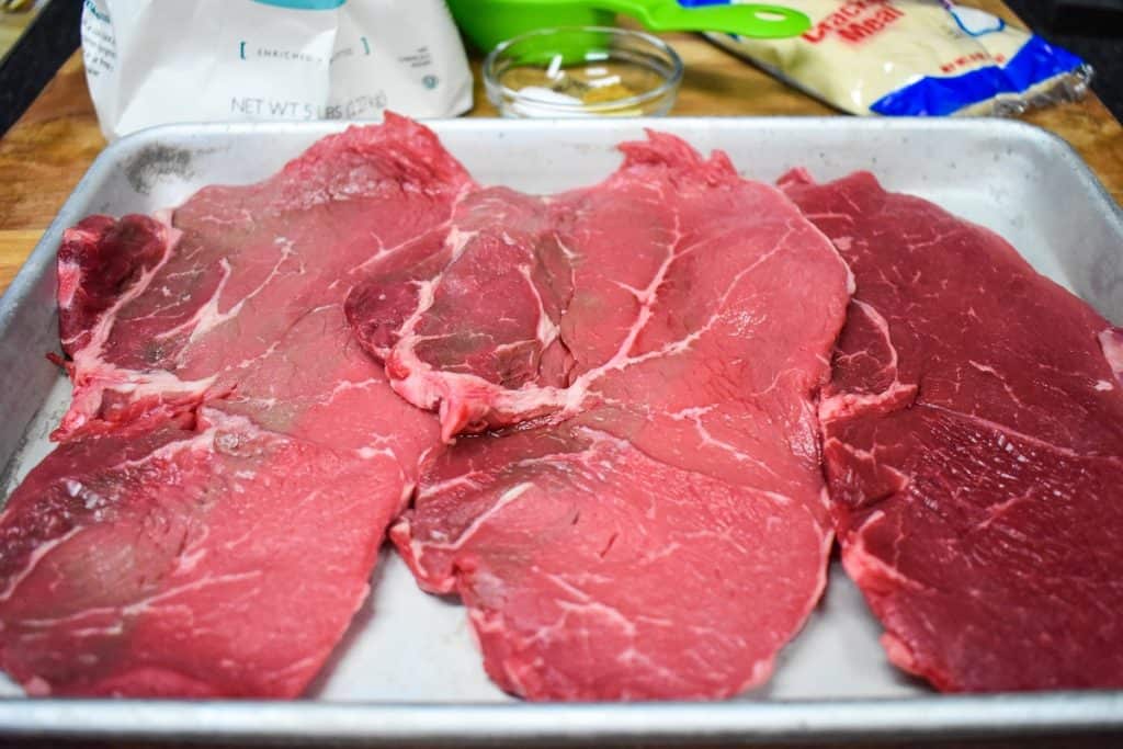 Three, large, raw palomilla steaks on a metal baking sheet.