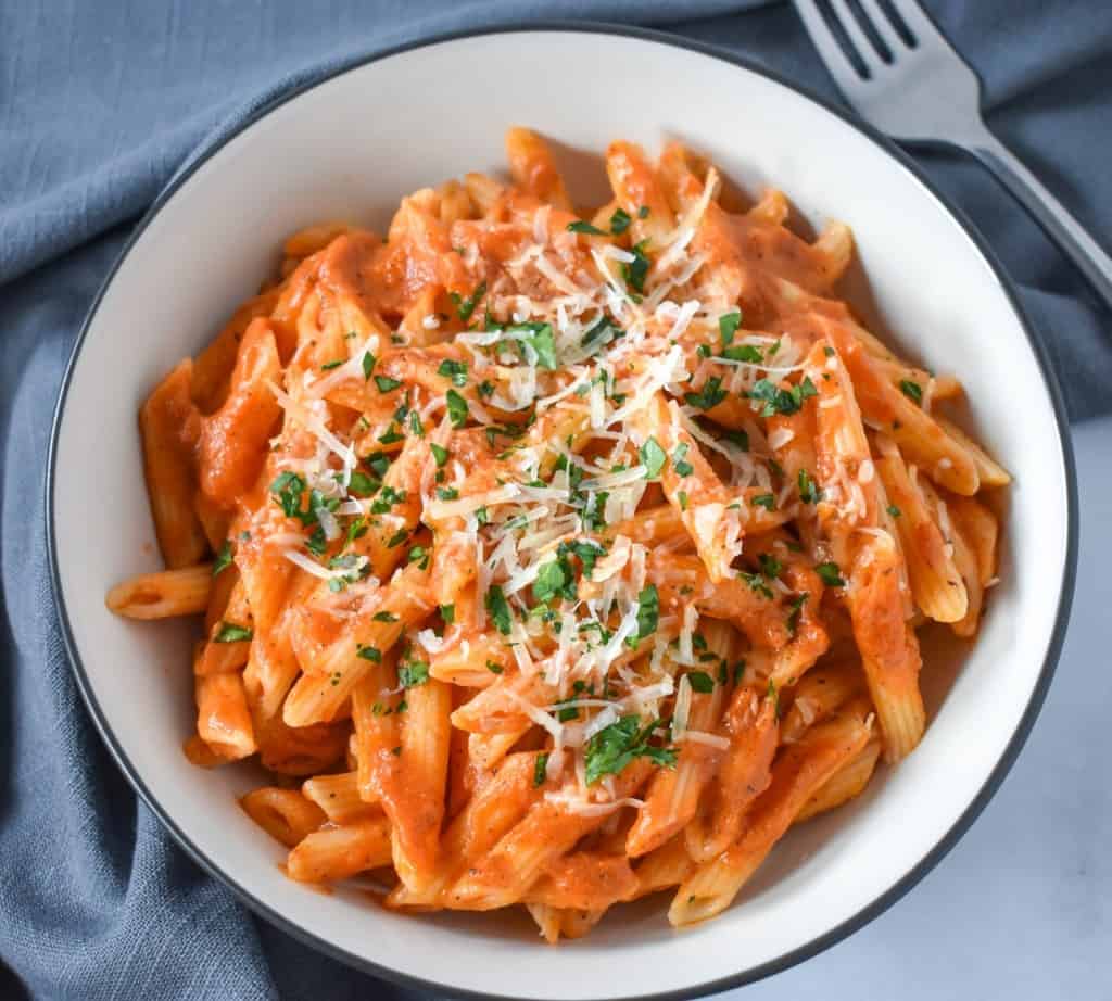Pasta with vodka sauce garnished with chopped parsley and parmesan cheese, served in a white bowl with a black rim with a gray linen and a fork in the upper right hand side.