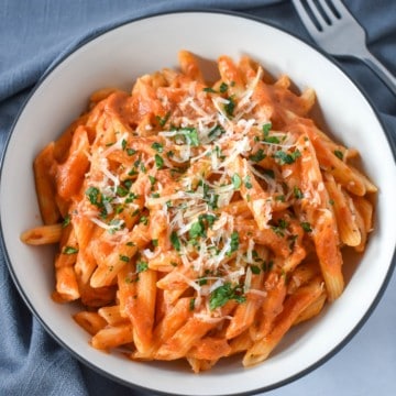 Pasta with vodka sauce garnished with chopped parsley and parmesan cheese, served in a white bowl with a black rim with a gray linen and a fork in the upper right hand side.