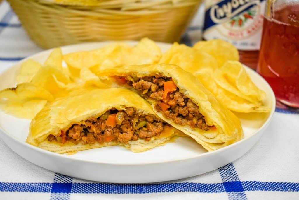 A ground beef stuffed biscuit, cut in half and arranged on a white plate with potato chips. In the background there is a weaved basket a bottle of hot sauce and a glass of punch.