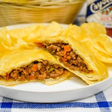 A ground beef stuffed biscuit, cut in half and arranged on a white plate with potato chips. In the background there is a weaved basket a bottle of hot sauce and a glass of punch.