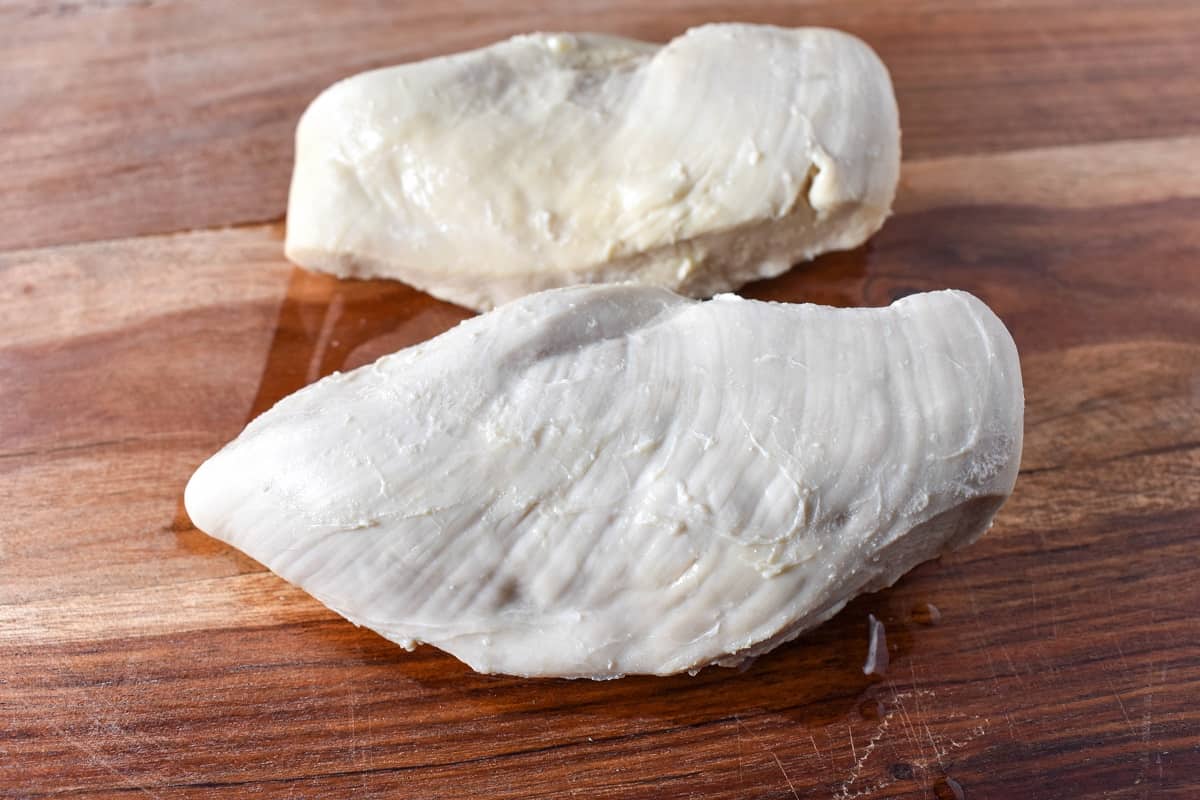 Two poached chicken breasts on a wood cutting board.