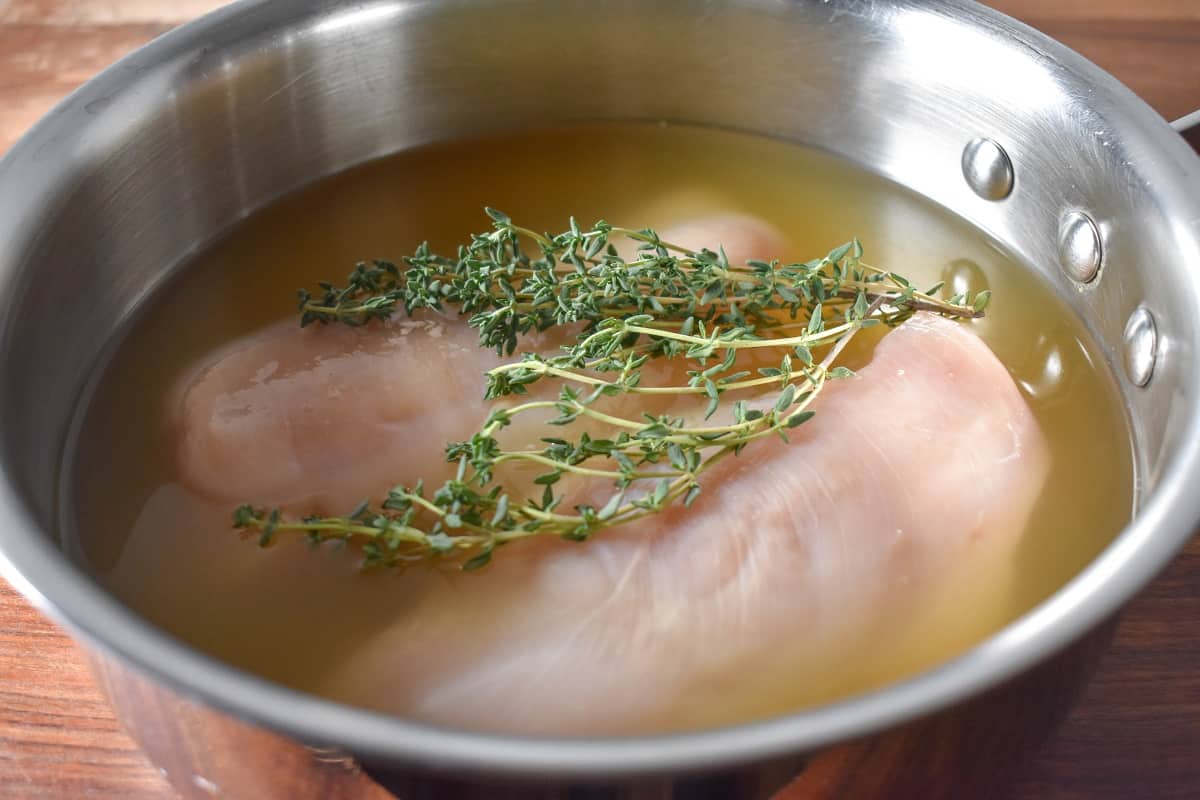 Two raw chicken breasts in a saucepan covered with chicken broth and thyme sprigs.