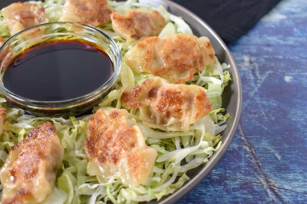 Pork potstickers arranged on a bed of shredded lettuce on a gray plate with a small bowl of soy sauce in the middle.