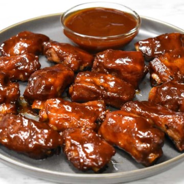 Pork riblets arranged on a large gray plate with a small bowl of barbecue sauce.