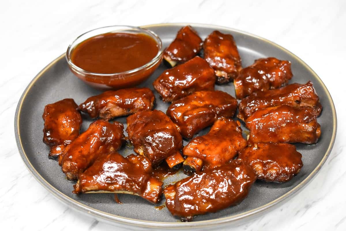 Pork riblets arranged on a large gray plate with a small bowl of barbecue sauce.