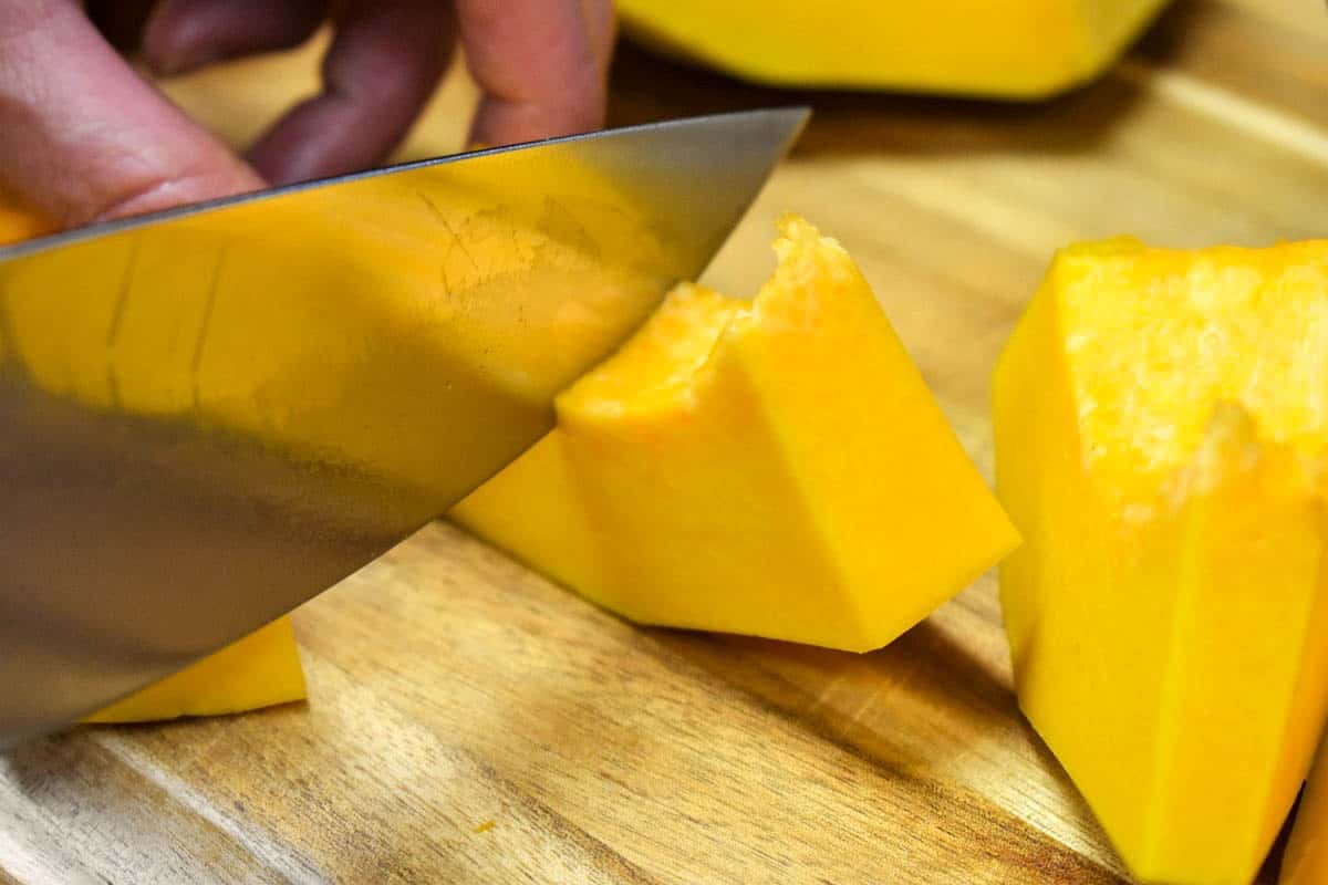 A peeled pumpkin chunk being cut into one to two inch pieces with a large knife.