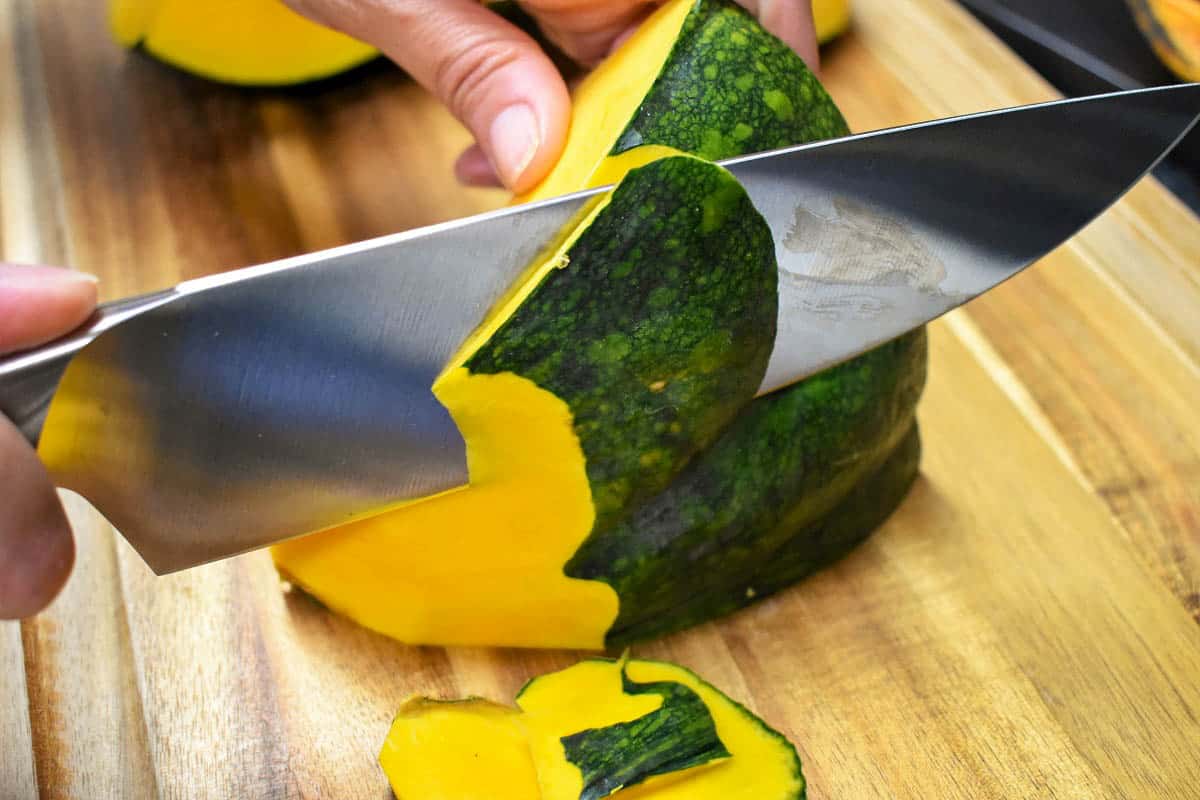 A chunk of pumpkin on a cutting board with a large knife cutting a thin piece of the peel off.