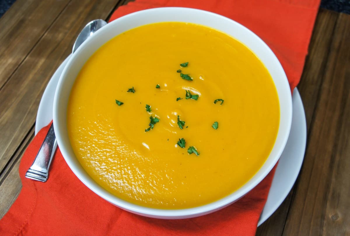 A top shot of the soup in a large white bowl set on an orange linen on a white plate with a spoon to the left.